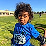 Malakai is registered to the contest to win money with this photo: child, boy, curly_hair, blue_shirt, graphic_tee, outdoor, grass, sunlight, squinting, playful, stroller, wagon, park, daytime, casual_clothing, smile, person, background, building, nature