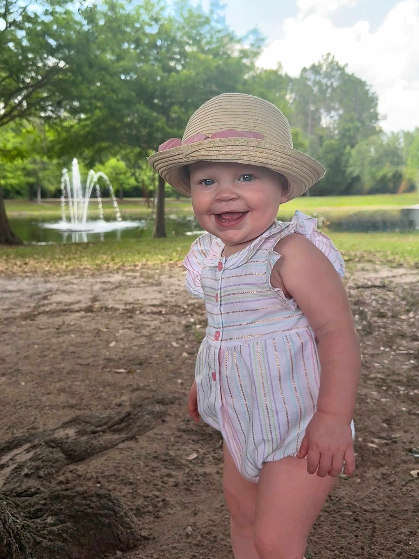 Gwendalyn joined the competition — help win amazing prizes! baby, child, smiling, hat, romper, outdoor, park, fountain, grass, tree, sunny, nature, happy, playful, person, summer, daylight, cute, portrait, standing