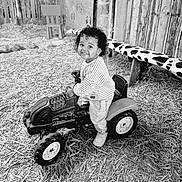 Naya a rejoint le concours — aidez-le/la à gagner de superbes lots ! toddler, toy_tractor, smile, curly_hair, outdoor, straw, wooden_fence, bench, cow_print, small_chair, child, play, happy, rustic, farm, person, jeans, striped_shirt, footwear, black_and_white
