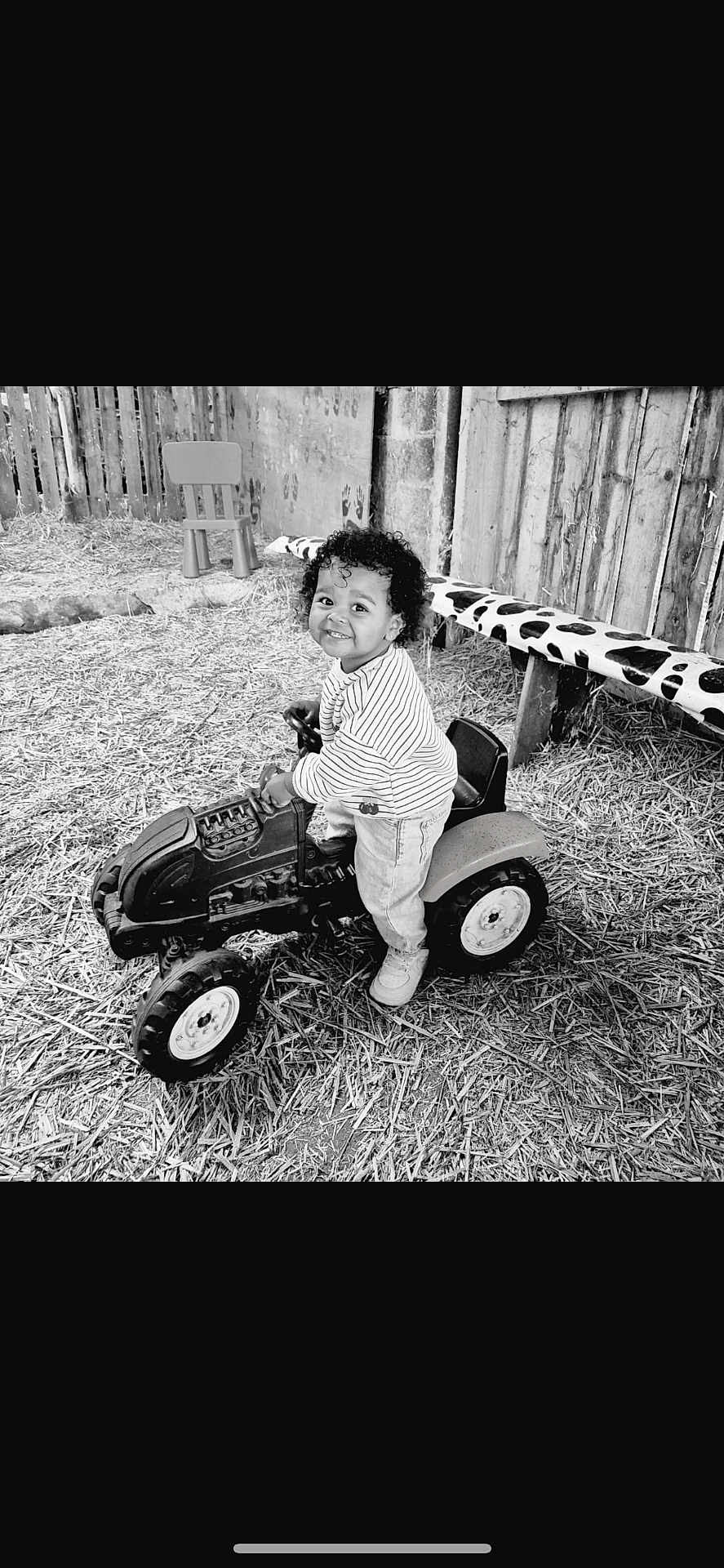 Naya a rejoint le concours — aidez-le/la à gagner de superbes lots ! toddler, toy_tractor, smile, curly_hair, outdoor, straw, wooden_fence, bench, cow_print, small_chair, child, play, happy, rustic, farm, person, jeans, striped_shirt, footwear, black_and_white