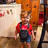barefoot, child, cute, denim_overalls, freezer, furniture, home, indoor, kitchen, person, pigtails, playing, red_shirt, sippy_cup, smiling, stickers, toddler, wood_paneling, wooden_floor, young_child