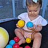 toddler, child, baby, ball, colorful_balls, trampoline, white_bow, white_top, puffy_sleeves, outdoor, backyard, netting, sitting, curious, playing, barefoot, pink_diaper, toy, daylight, young_child