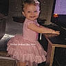 toddler, girl, pink_dress, bow, smiling, barefoot, computer, desk, indoor, floor_tiles, furniture, child, portrait, cute, standing, happy, technology, room, lighting, text_overlay