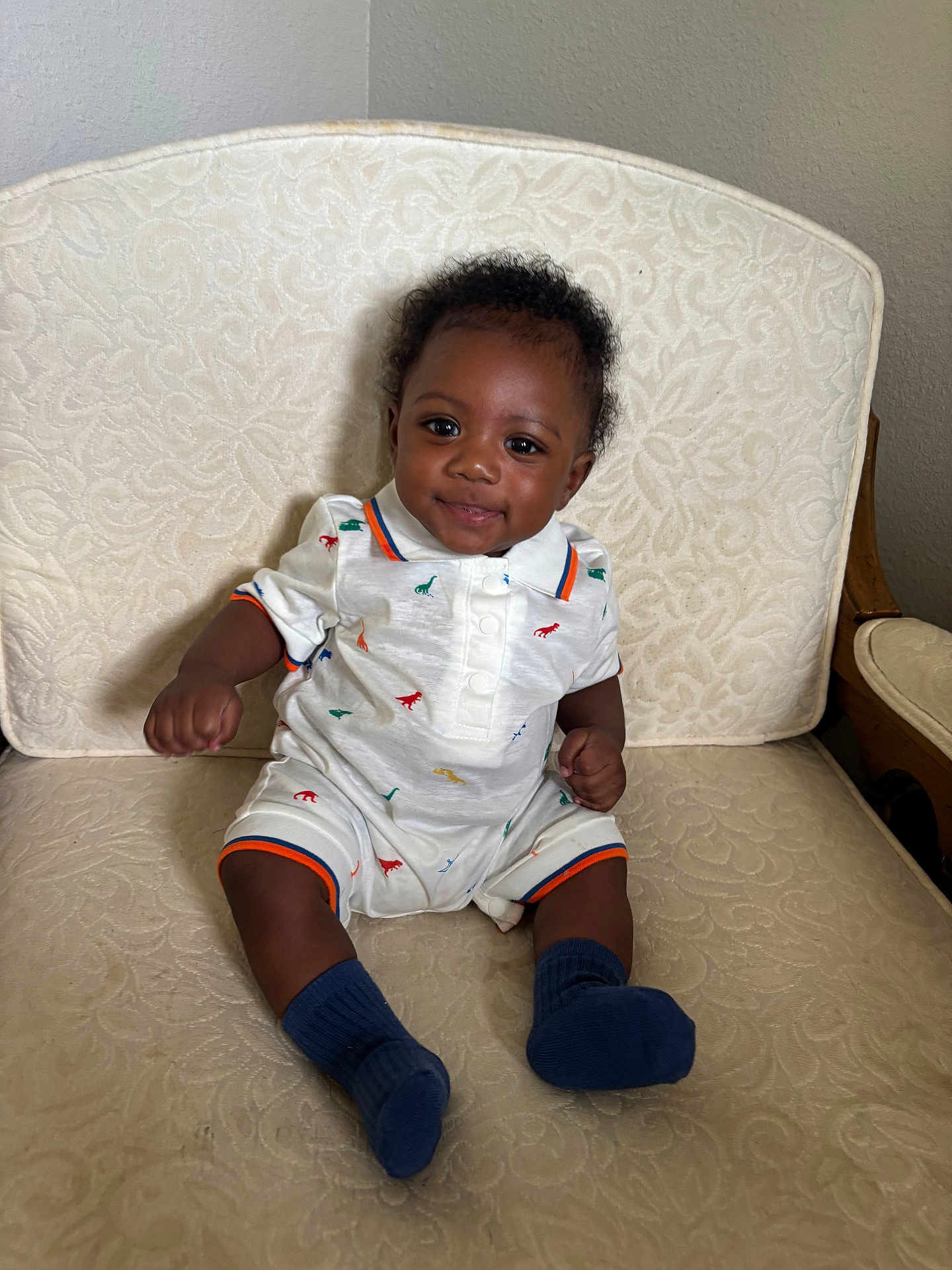 Raziel joined the competition — help win amazing prizes! baby, child, smiling, sitting, indoor, clothing, dinosaur_print, socks, chair, patterned_fabric, cute, happy, portrait, infant, toddler, person, furniture, dark_skin, curly_hair, casual
