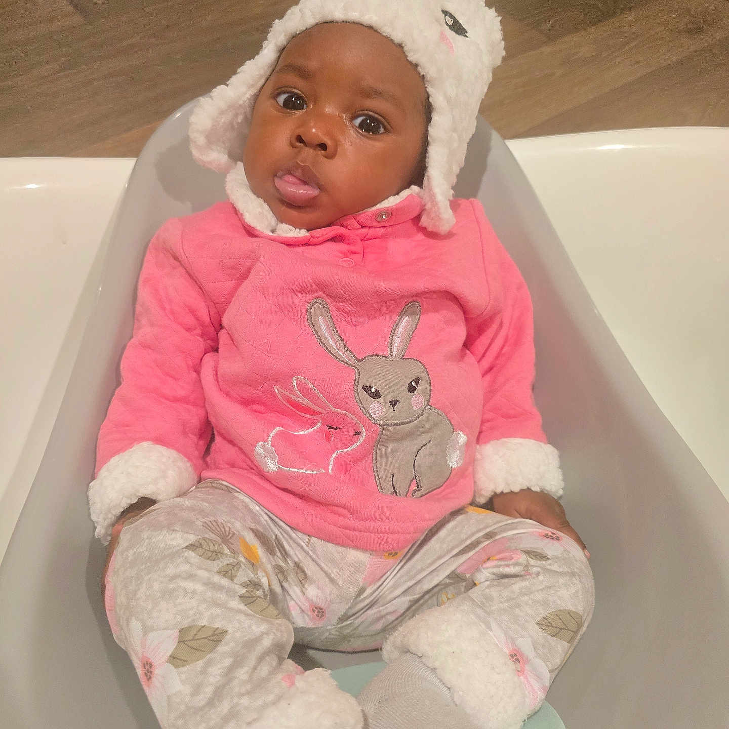 Ayla joined the competition — help win amazing prizes! adorable, baby, bunny_design, child, cozy, cute_expression, eyes, face, fluffy_hat, indoor, infant, person, pink_sweater, portrait, sitting, socks, tongue_out, warm_clothing, white_tub, wooden_floor