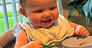 Kinsley joined the competition — help win amazing prizes! baby, bib, bowl, child, cute, feeding, food, green_plants, happy, high_chair, home, indoor, infant, meal, person, smiling, spoon, table, vertical_blinds, window