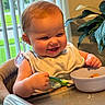 baby, bib, bowl, child, cute, feeding, food, green_plants, happy, high_chair, home, indoor, infant, meal, person, smiling, spoon, table, vertical_blinds, window