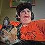 animal, black_fur, brown_fur, calico_cat, cap, cat, close_up, curious, face, feline, green_eyes, headphones, indoor, orange_shirt, painting, person, pet, portrait, selfie, wall