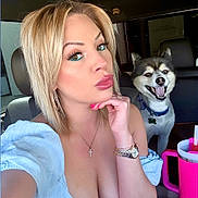 Loki Papi Bear is registered to the contest to win money with this photo: person, dog, pet, selfie, car_interior, front_seat, blonde_hair, makeup, jewelry, necklace, watch, tumbler, pink_tumbler, smiling_dog, open_mouth, pouting, low_cut_top, headrest, accessory, companion