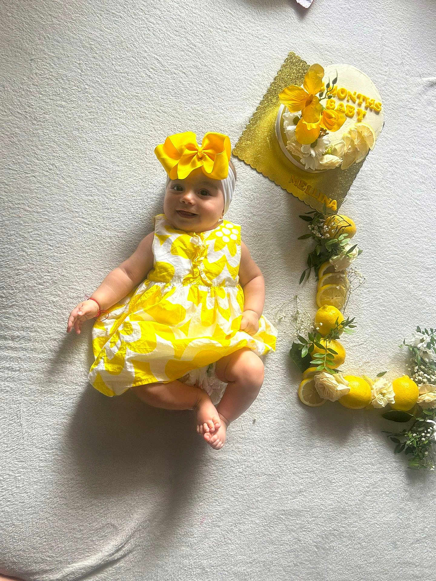 Mellina participe au concours pour gagner de l'argent avec cette photo : 4_months, baby, blanket, bow, cake, celebration, child, cute, decorations, flowers, happy, headband, indoors, infant, laying_down, lemons, milestone, portrait, smiling, yellow_dress