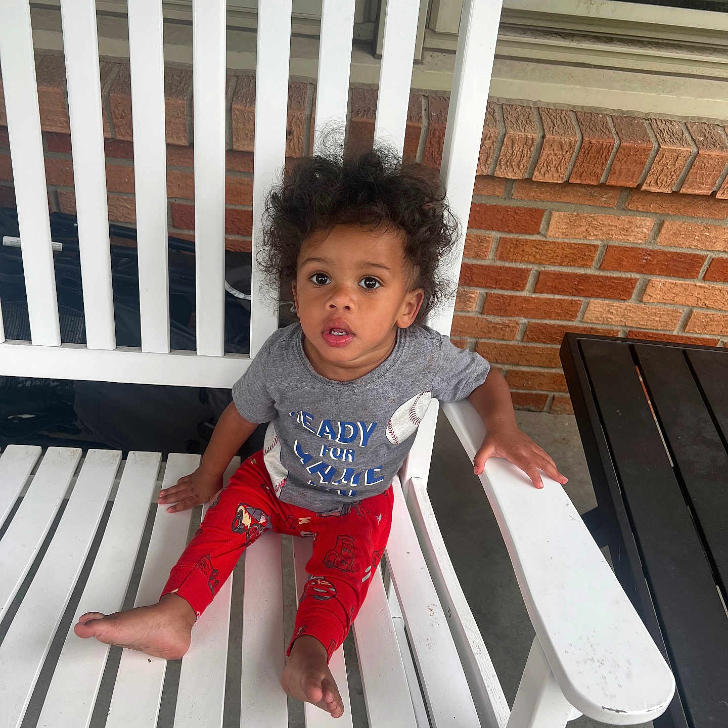 Dorion joined the competition — help win amazing prizes! barefoot, black_table, brick_wall, cartoon_prints, casual_clothing, child, curly_hair, expression, grey_tshirt, hand_on_armrest, indoor_outdoor, looking_up, porch, red_pants, sitting, toddler, white_chair, wide_eyes, wooden_slats, young_child