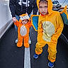 adult, asphalt, baby, car, child, costume, cute, holding_hands, hood, hoodie, jeans, line, orange, outdoor, parking_lot, person, playful, shoes, smile, wings