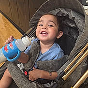 Alex is registered to the contest to win money with this photo: baby, baby_bottle, black_padded_wall, blue_shirt, child, clothing, face, hand, happy, head, indoor, person, seatbelt, smiling, stroller, temporary_tattoo, toddler, toy, wheels, wooden_floor