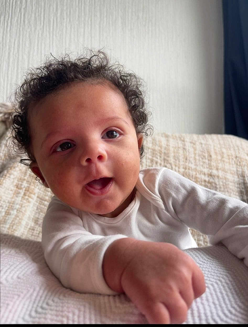 Ellis is registered to the contest to win money with this photo: baby, child, close_up, couch, curiosity, curly_hair, cute, expression, face, hand, happy, indoor, infant, person, portrait, skin, smiling, texture, white_clothing, young_child