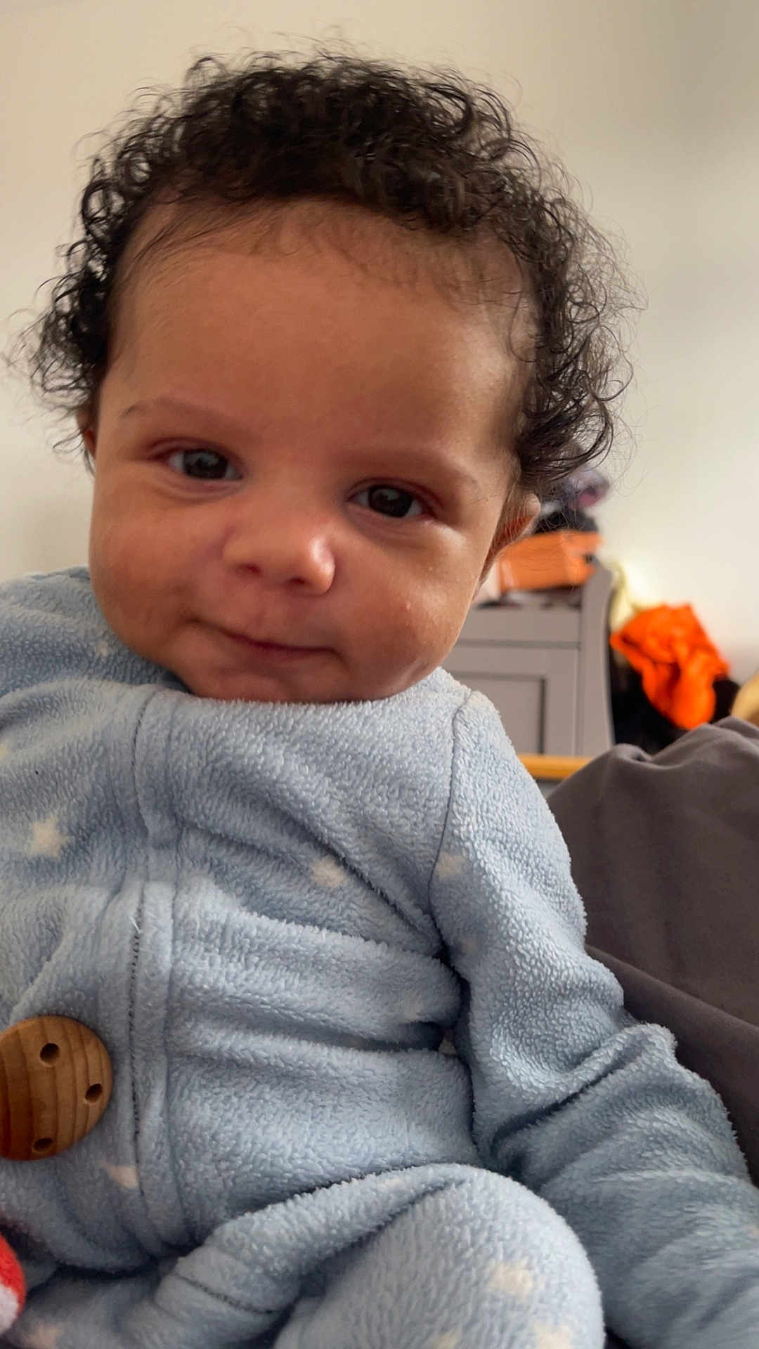 Ellis joined the competition — help win amazing prizes! baby, child, curly_hair, blue_onesie, indoor, soft_texture, face, portrait, clothing, seated, infant, cute, young_child, expression, looking_at_camera, warm_clothing, home, close_up, person, adorable