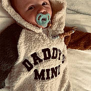Keenan is registered to the contest to win money with this photo: baby, child, pacifier, hooded_onesie, text_on_clothing, fluffy_texture, blanket, soft_fabric, infant, cute, sleepwear, lying_down, indoors, warm_clothing, newborn, face, head, person, comfort, expression