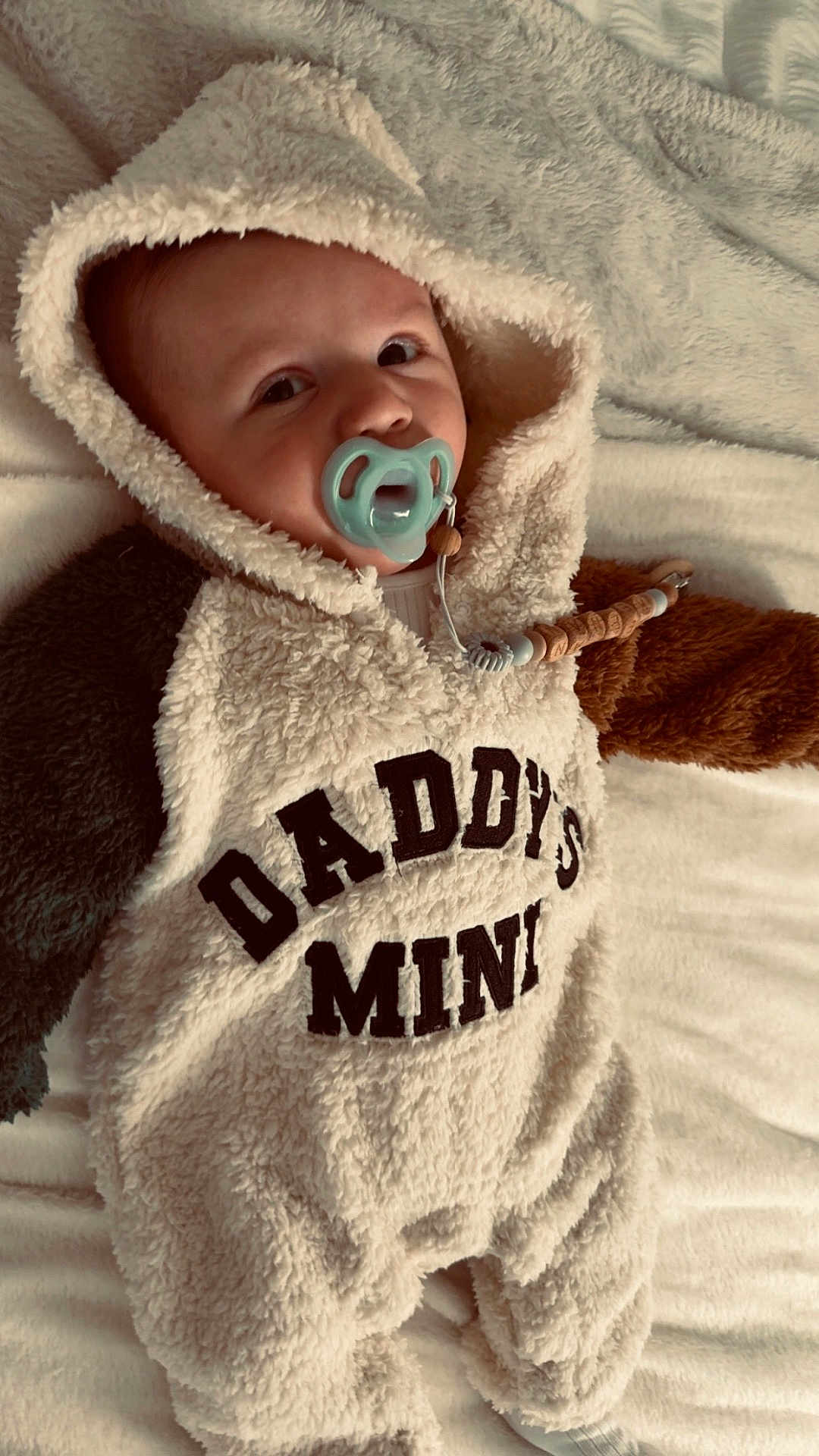 Keenan is registered to the contest to win money with this photo: baby, child, pacifier, hooded_onesie, text_on_clothing, fluffy_texture, blanket, soft_fabric, infant, cute, sleepwear, lying_down, indoors, warm_clothing, newborn, face, head, person, comfort, expression