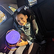 Brooklynn is registered to the contest to win money with this photo: child, car_seat, smiling, seatbelt, purple_container, sunlight, car_interior, window, happy, young_person, clothing, portrait, seat, daylight, safety, child_safety, vehicle, casual, childhood, inside_car