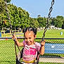child, swing, playground, smile, happy, outdoor, grass, trees, sunny, blue_sky, fence, footwear, pink_shirt, braided_hair, young_girl, park, summer, daylight, joyful, recreation
