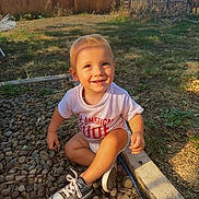 Rocky is registered to the contest to win money with this photo: toddler, child, smiling, sitting, rocks, backyard, sunlight, grass, fence, sneakers, clothing, happy, outdoor, person, nature, wood, daylight, casual, cute, playing