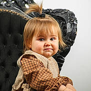 Enola participe au concours pour gagner de l'argent avec cette photo : toddler, child, portrait, smile, face, hair, pony_tail, clothing, vest, checkered, chair, black_chair, furniture, indoor, cute, expression, person, baby, sitting, young_child