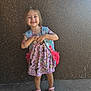 child, girl, smiling, backpack, dress, princesses, sandals, bracelet, standing, concrete_floor, wall, sunlight, happy, outdoor, portrait, cute, fashion, person, footwear, accessory