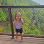 child, girl, outdoor, forest, wooden_deck, metal_fence, nature, smiling, summer, happy, standing, blue_shorts, pink_sandals, trees, greenery, daylight, scenic, casual_clothing, portrait, young