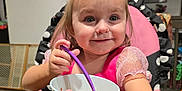 Freya is registered to the contest to win money with this photo: toddler, child, smiling, eating, bowl, spoon, high_chair, indoor, table, pink_clothing, face, happy, person, hair, room, painting, wooden_table, polka_dot, sitting, cute