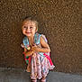 child, girl, smiling, backpack, dress, sandals, standing, blue_eyes, outdoor, concrete_floor, textured_wall, happy, cute, portrait, person, summer_clothing, footwear, young, innocent, playful