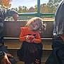 child, girl, smiling, orange_clothing, black_skirt, pumpkin_design, apple, bench, outdoor, autumn, trees, sunlight, people, casual_clothing, happy, portrait, festive, family, nature, daylight