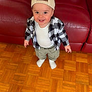 Ayaan a rejoint le concours — aidez-le/la à gagner de superbes lots ! backwards_cap, cap, casual_clothing, checkered_shirt, child, cute, happy, home, indoor, khaki_pants, parquet_floor, person, portrait, red_couch, smiling, socks, standing, toddler, white_tshirt, wooden_floor
