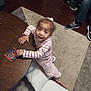toddler, child, table, wooden_table, striped_shirt, pink_pants, remote_control, bowl, nuts, carpet, sneakers, living_room, person, floor, toy, curly_hair, white_bow, indoor, cushion, blur