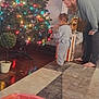 toddler, adult, christmas_tree, ornament, lights, pajamas, indoor, holiday, decorating, family, carpet, wood_floor, potted_plant, basket, barefoot, beard, child, adult_male, home, festive