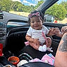 baby, infant, car_interior, child, smiling, headband, white_clothing, hand_holding, person, seat, window, trees, daytime, sunny, clothing_accessory, tattoo, food_container, car_dashboard, seatbelt, blanket