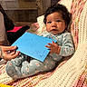 Azorie is registered to the contest to win money with this photo: baby, infant, child, curly_hair, blue_pajamas, floral_pattern, knit_blanket, envelope, handwritten_note, tattooed_hand, children_book, indoor, cozy, cute, person, sitting, portrait, soft_lighting, warm_colors, expression