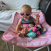 Nicole is registered to the contest to win money with this photo: baby, child, infant, sunglasses, heart_shaped_sunglasses, pink, baby_chair, seated, toys, stuffed_animals, playroom, indoor, couch, blanket, carpet, striped_clothing, smiling, portrait, fashion, cute