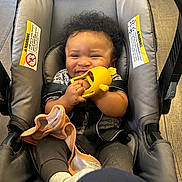 Seven is registered to the contest to win money with this photo: baby, car_seat, child, toy, smile, curly_hair, clothing, fabric, seatbelt, infant, happy, indoor, floor, hands, blanket, person, sitting, wood_floor, warning_label, cute