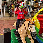 Sebastian is registered to the contest to win money with this photo: child, boy, mechanical_horse, toy_ride, red_hoodie, ole_miss, elf_hat, smiling, playful, shoe_store, storefront, glass_windows, reflection, footwear, sneakers, indoor_outdoor, floor_tiles, colorful, fun, portrait