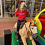 child, boy, mechanical_horse, toy_ride, red_hoodie, ole_miss, elf_hat, smiling, playful, shoe_store, storefront, glass_windows, reflection, footwear, sneakers, indoor_outdoor, floor_tiles, colorful, fun, portrait