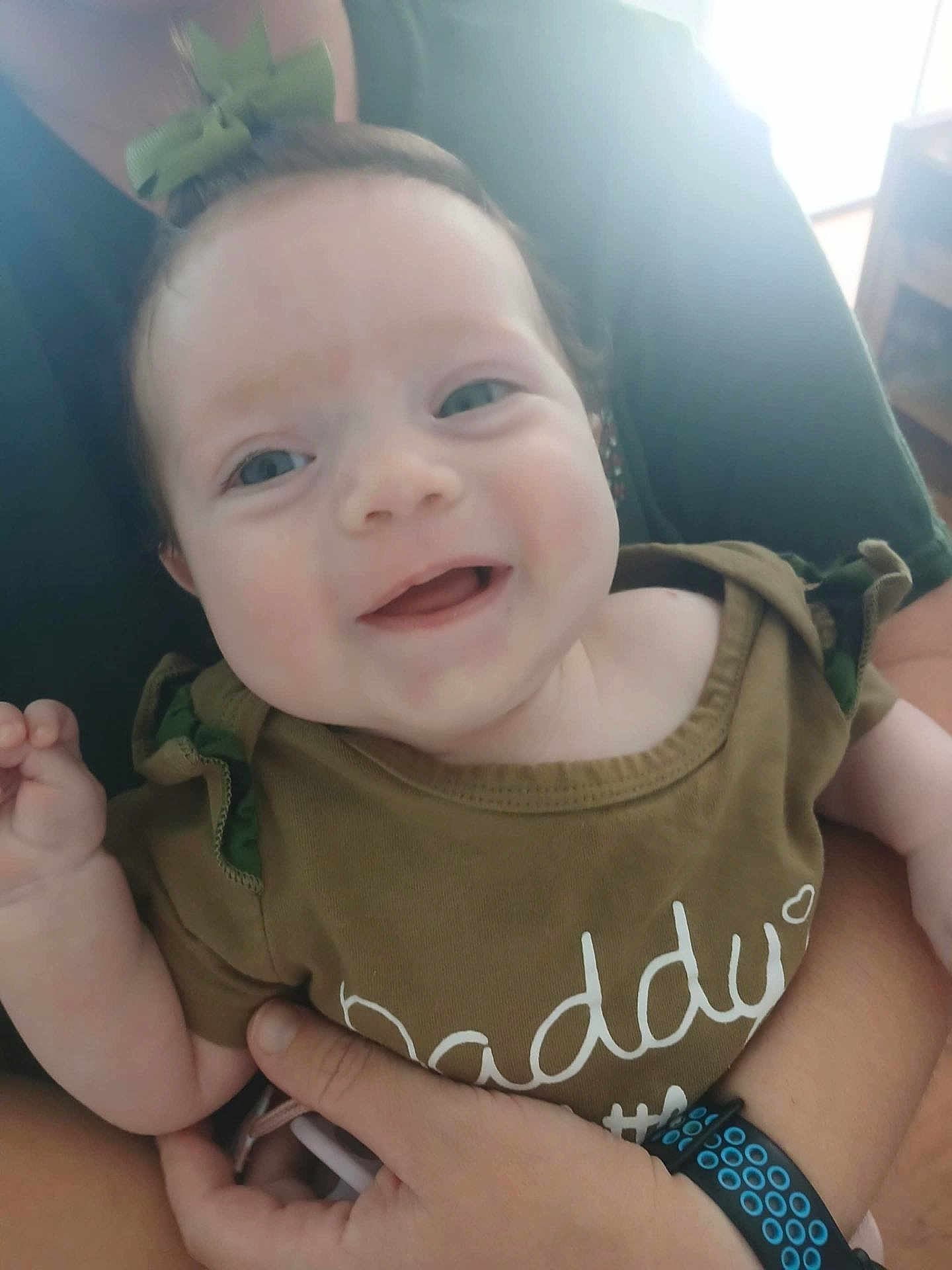 Mattalyn is registered to the contest to win money with this photo: baby, child, smiling, green_bow, green_clothing, adult_hand, wristband, indoor, portrait, face, happy, infant, close_up, person, casual, holding, cute, young_child, apparel, human