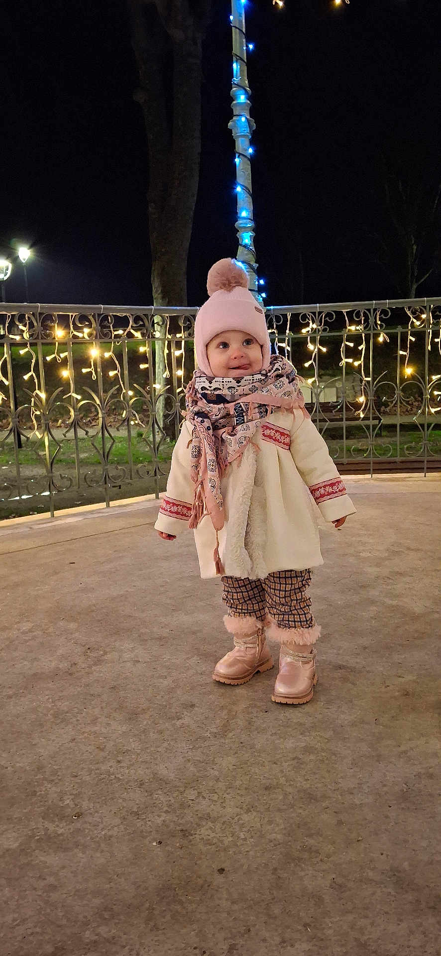 Alexia participe au concours pour gagner de l'argent avec cette photo : toddler, child, winter_clothing, hat, scarf, boots, outdoor, night, lights, fairy_lights, pole, fence, curious, smile, cold_weather, park, concrete_floor, cute, fashion, cozy