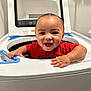 baby, child, washing_machine, laundry, smiling, happy, red_shirt, indoor, face, hands, appliance, home, cute, playful, infant, person, head, expression, cleaning, household