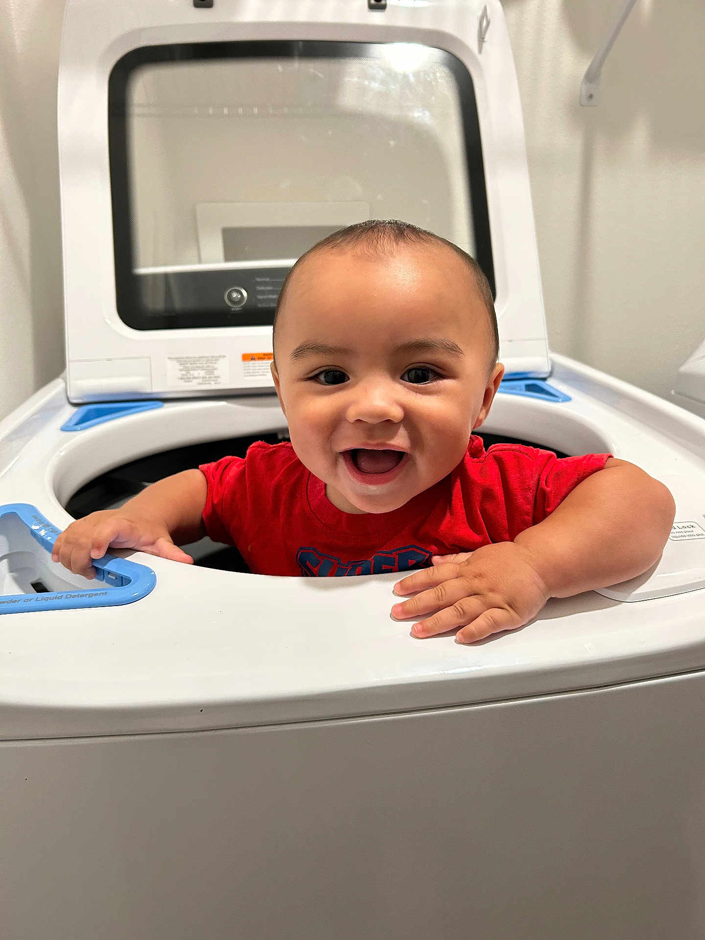 Wes joined the competition — help win amazing prizes! baby, child, washing_machine, laundry, smiling, happy, red_shirt, indoor, face, hands, appliance, home, cute, playful, infant, person, head, expression, cleaning, household