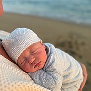 Brooks joined the competition — help win amazing prizes! baby, newborn, sleeping, infant, white_hat, white_clothing, beach, ocean, blurred_background, close_up, peaceful, soft_texture, adult, holding, cuddling, warm, tender_moment, portrait, outdoor, nature