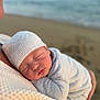 baby, newborn, sleeping, infant, white_hat, white_clothing, beach, ocean, blurred_background, close_up, peaceful, soft_texture, adult, holding, cuddling, warm, tender_moment, portrait, outdoor, nature