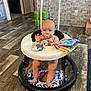baby, walker, pacifier, toy, stuffed_animal, floor, wood_floor, brick_wall, indoor, child, colorful, book, hand, face, cushion, person, sitting, cute, infant, home
