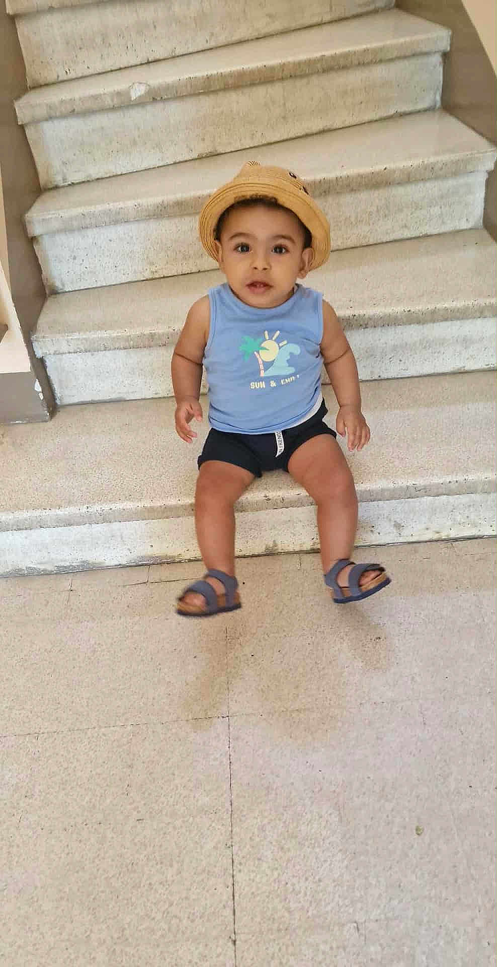 Raheem participe au concours pour gagner de l'argent avec cette photo : toddler, child, hat, stairs, sitting, blue_shirt, shorts, sandals, curious, indoor, concrete, floor, person, portrait, casual_clothing, young_child, expression, looking_up, bare_arms, cute