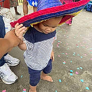 Raphaël a rejoint le concours — aidez-le/la à gagner de superbes lots ! barefoot, casual_clothing, celebration, child, colorful, concrete_floor, confetti, festival, footwear, hand_holding, hat, legs, outdoor, people, person, shorts, sneakers, sombrero, striped_shirt, toddler