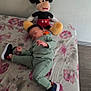 bed, bedspread, child, cute, floor, floral_pattern, furniture, green_clothing, home, indoor, mickey_mouse, motion_blur, person, playful, plush_toy, sneakers, soft_toy, toddler, toy, wall