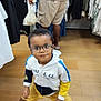 adult, boy, casual_clothing, child, clothing_store, curious, floor, glasses, hanger, hoodie, indoor, person, pose, shopping, sleeves, sneakers, standing, store, wooden_floor, yellow_shorts
